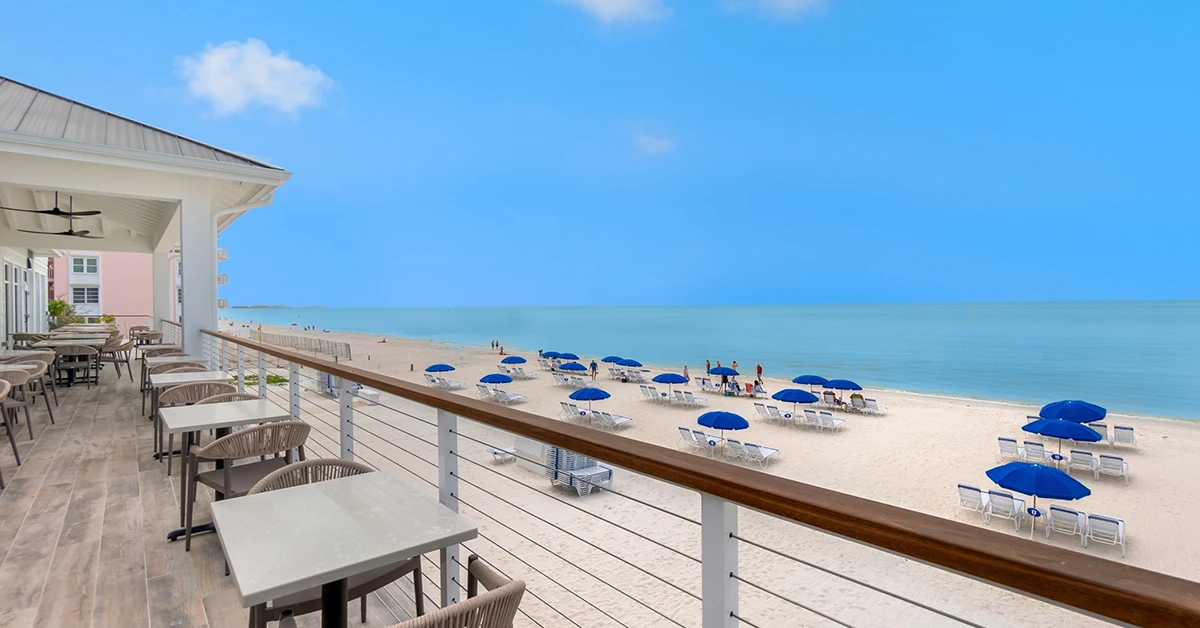 Patio with tables overlooking sandy beach and blue ocean with umbrellas.