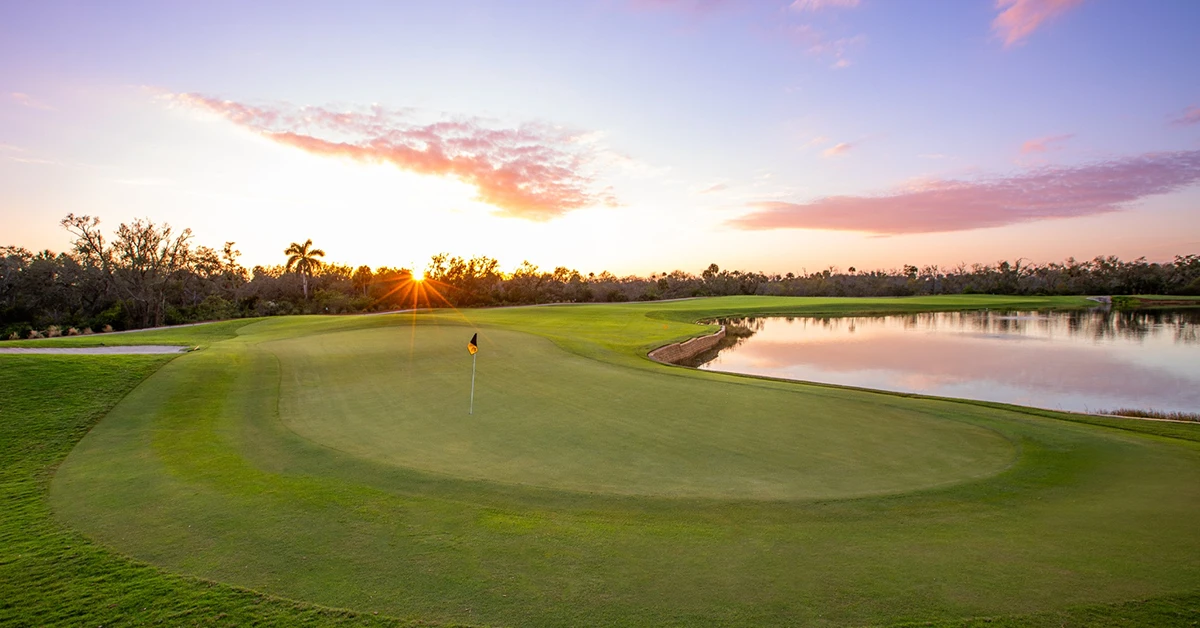 Golf course green with flag beside water under sunrise sky.