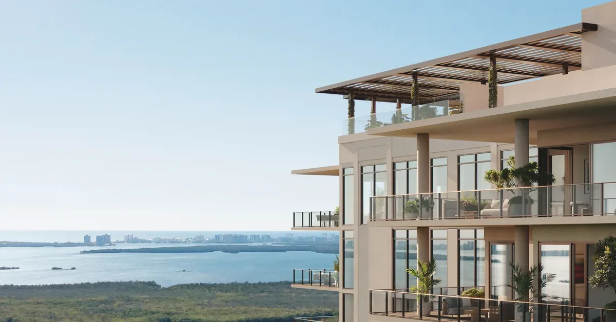 Luxury waterfront condominium balconies with glass railings overlook lush mangroves and a shimmering bay with a city skyline in the distance.